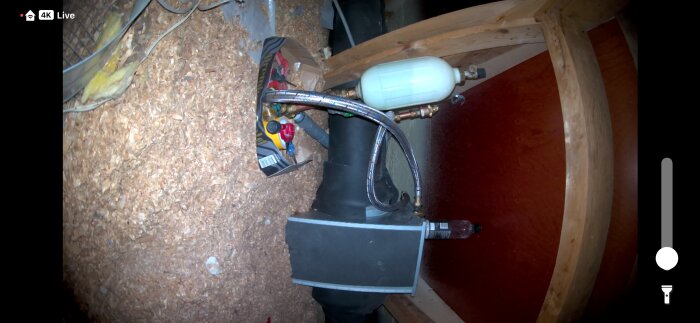 Anslutningar och rör kopplade till ett frikylesystem i ett trångt utrymme, med isoleringsmaterial och trästruktur synlig i bakgrunden.