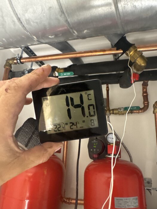 Termometer visar 14°C, monterad nära rör och ventiler i ett frikylesystem. En hand håller mätaren, omgiven av rör och röda cylindrar.