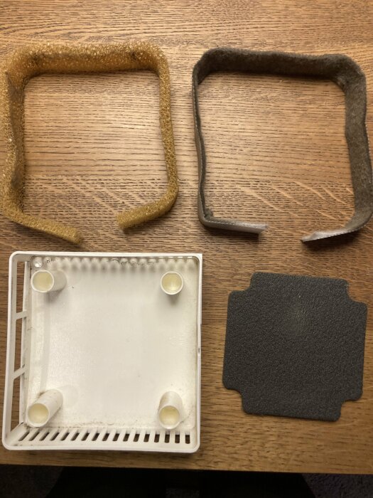 Tre fyrkantiga filter av olika material och en vit plastdel, placerade på ett trägolv. Ett filter verkar vara ett svart kondensfilter.