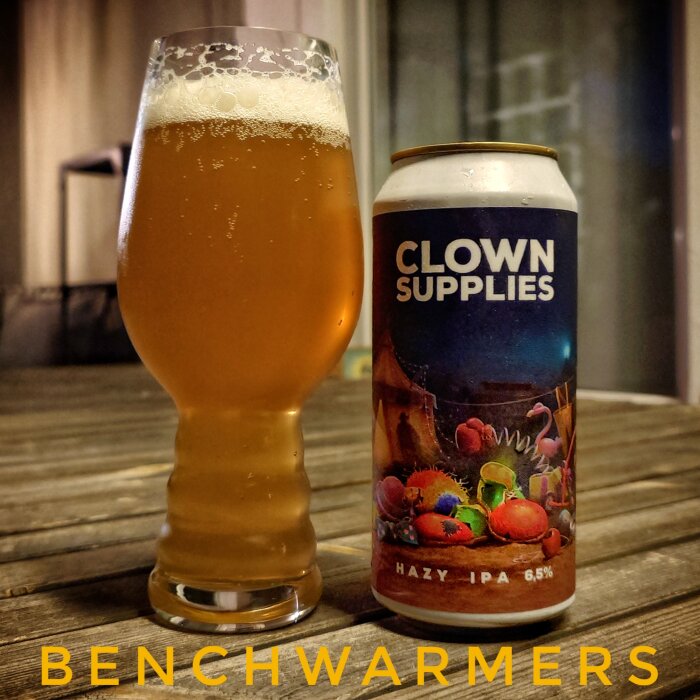 Ölglas och burk med etiketten "Clown Supplies" Hazy IPA 6,5% på ett träbord.