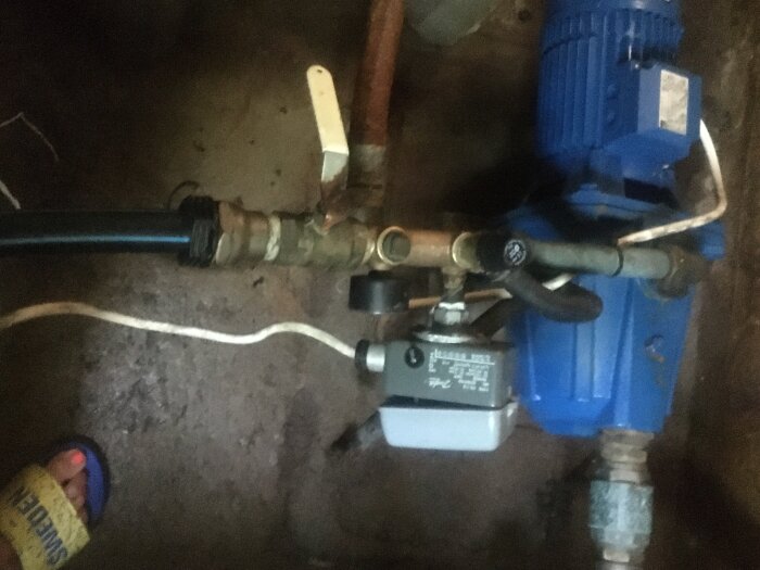 Vattenpump i pumphus med motor och röranslutningar, inklusive en ventil och slangar, på ett betonggolv.