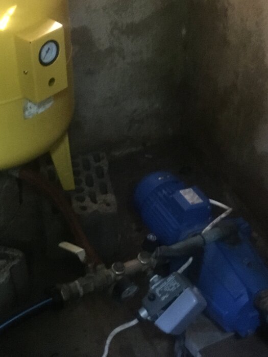 Ett pumphus med en blå vattenpump och en gul tryckbehållare, samt olika ventiler och rör kopplade till systemet.