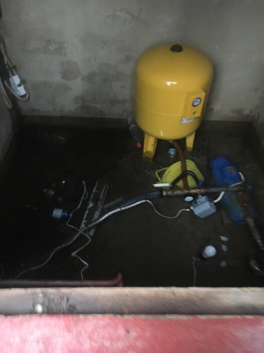 Källare med gammal vattentank och ledningar, slarvigt placerade slangar och elkontakter på golvet, med en gul tank i hörnet.