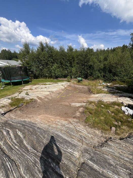 Utgrävt område på ett berg med markeringar för en stenläggning, omgiven av grönska och en gräsmatta med en studsmatta och skottkärra.