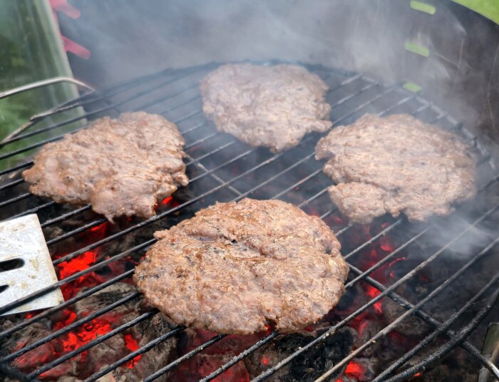 Fyra saftiga nötfärsburgare grillas på en utegrill med glödande kol och rök.