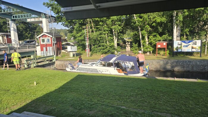 Båt vid Dalslands kanal med personer ombord, omgiven av grönska och skyltar. Solig dag med kanalområde i bakgrunden.