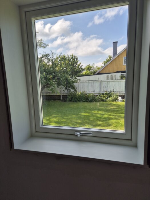 Utsikt från ett fönster med vitmålad fönsterbräda i obehandlad furu, ser ut mot en grön gräsmatta och ett hus i bakgrunden.