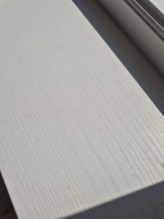 Nymålad fönsterbräda i obehandlat furu där träådringen syns genom vit färg. Solbelyst yta visar texturen och detaljer i träet.