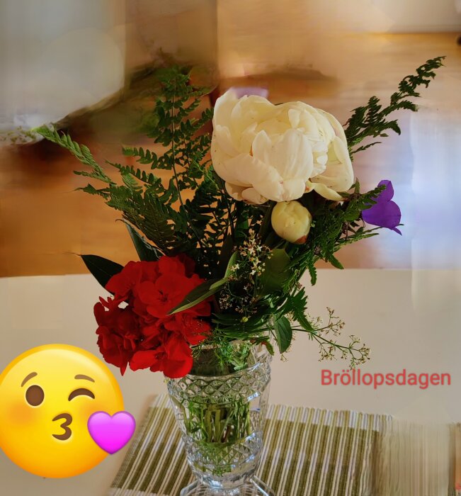 Blombukett i en glasvas på ett bord, med vita och röda blommor samt gröna blad. Texten "Bröllopsdagen" och en blinkande emoji syns.