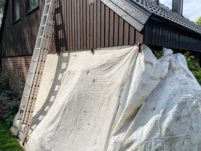 Stege lutad mot träfasad, täckt med presenning inför målning.