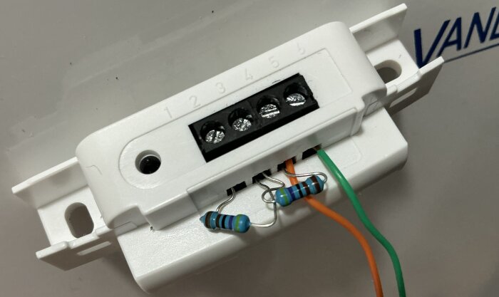 Elektrisk kopplingsbox med fyra portar, orange och grön ledning ansluten, två resistor-komponenter synliga, nummermärkning ovanför.