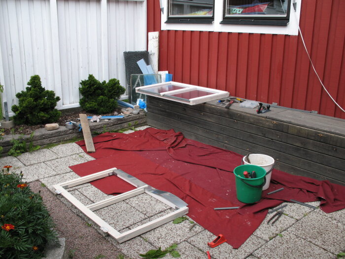 Renoveringsprojekt med fönsterram på uteplats, röda plattor, verktyg och hinkar. Rustik träbänk och grönska i bakgrunden.