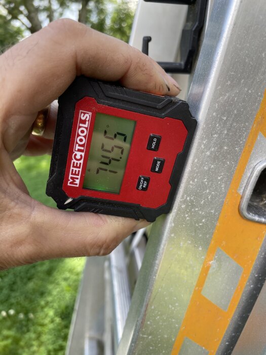 Digital vinkelmätare visar 74,5 grader mot en stege utomhus. En hand håller i verktyget.