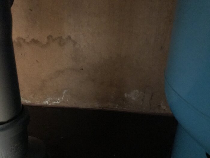 Vattenskada med svart missfärgning på köksvägg, synlig vid trasig plastmatta, indikeras av förhöjda fuktvärden och upphöjda golvbrädor.