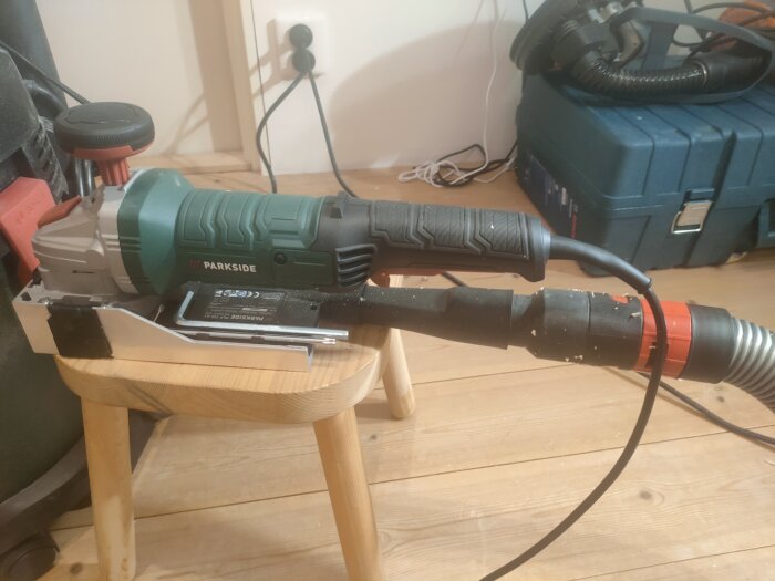 Parkside lackfräs placerad på en pall bredvid en blå verktygslåda, ansluten till en dammsugarslang i ett rum med trägolv.