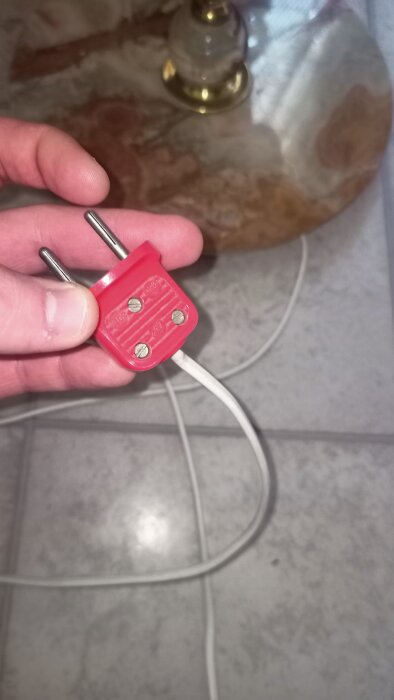 En hand håller i en röd stickkontakt med en vit kabel mot en marmorbordsskiva.