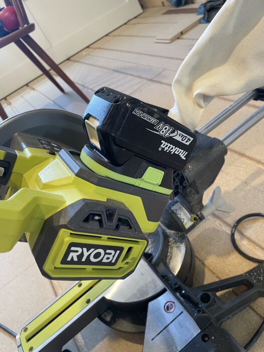 Gul och svart Ryobi kap- och geringssåg med Makita batteriadapter, placerad på golv i ett rum.