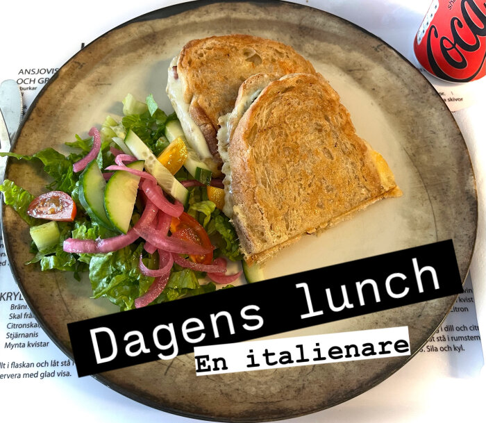 Grillad levainmacka med pesto, mozzarella, oliver, parmaskinka, serverad med sallad och picklad rödlök på tallrik, Coca-Cola-burk bredvid.