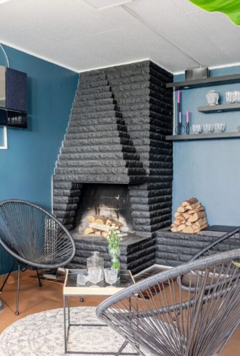 Öppen spis i ett poolrum med svart tegel, staplat ved, två fåtöljer och ett bord framför. Väggen är målad i blå nyans.