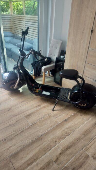 Elektrisk scooter inomhus, med synliga kablar och öppen panel, står på trägolv bredvid en stol och glasdörr.