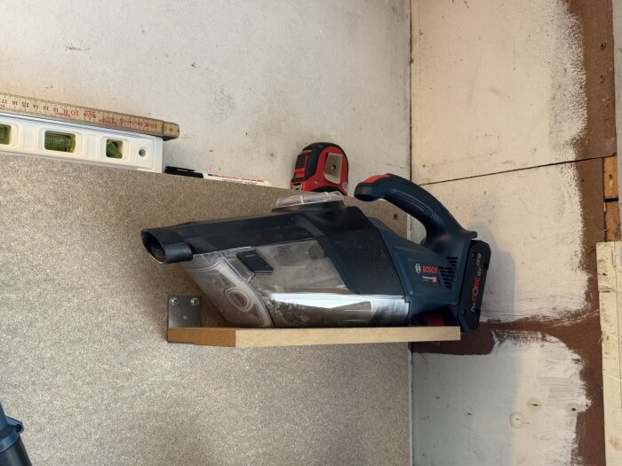 En hylla på väggen med en handhållen dammsugare, vattenpass och måttstock placerad ovanpå hyllan.