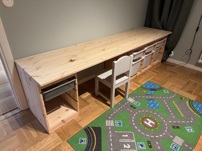 Ett träskrivbord med hyllor och plastlådor står mot en vägg. Framför står en liten stol och en färgglad lekmatta med vägmotiv.
