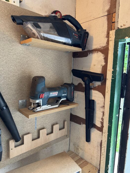 Verktygsförvaring med hyllor: en dammsugare överst, en såg i mitten och hållare för verktyg längst ner, vid en verkstadsvägg.