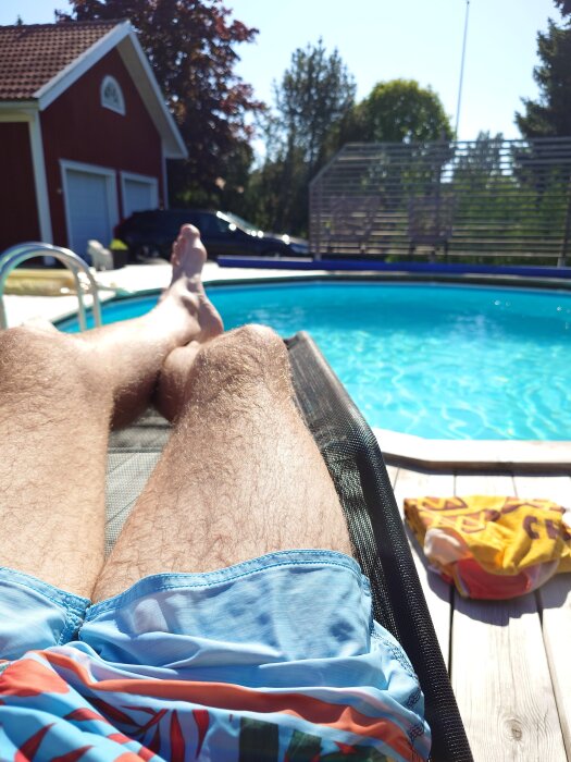 Person i solstol vid pool, benen i fokus, bär blå badbyxor. Rött hus och bil i bakgrunden, handduk på trädäck.