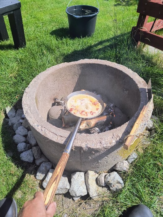 Utomhusgrilla vid en eldstad av cement och stenar, en hand håller en stekpanna med mat, omgiven av grön gräsmatta och en svart plasthink.