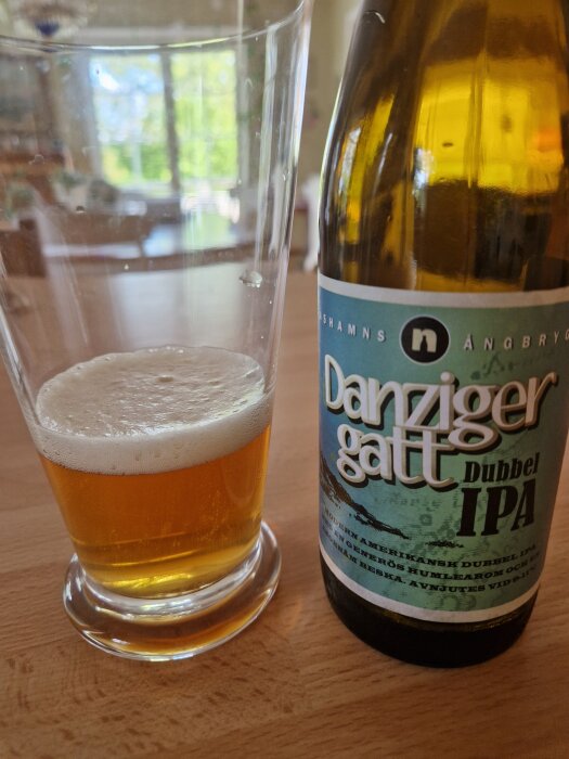 Flaska och glas med Danziger Gatt Dubbel IPA öl på ett bord i bakgrund av ett ljust rum.