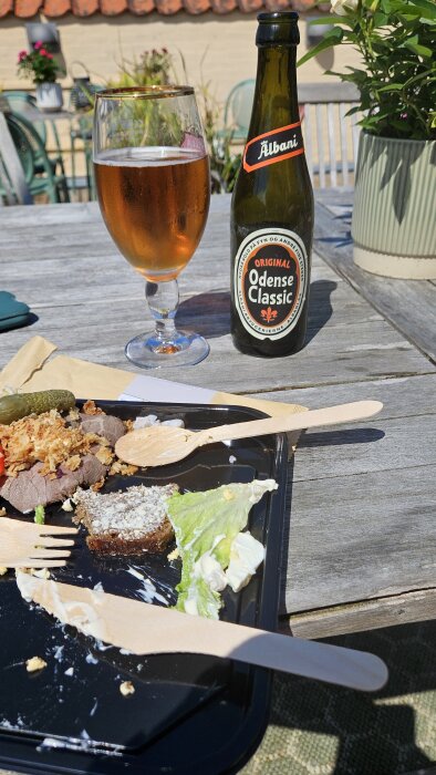 Flaska och glas med Odense Classic öl bredvid en tallrik med smørrebrød på ett utomhusbord.