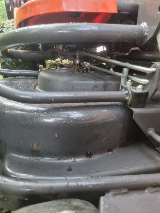 Närbild av mekaniken på en Husqvarna Rider 13, med fokusering på klippdäcket och dess fästen, delvis täckt med vattendroppar och gräs.