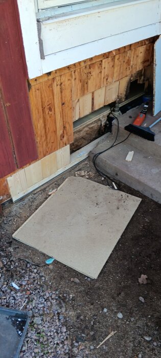 Vägg av trä med synlig skada vid basen, visar reparationsarbete vid grund; byggmaterial och verktyg synliga på marken.