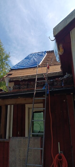 Takrenovering pågår med blå presenning täckande taket, stege lutad mot väggen, och takpannor staplade nedanför.