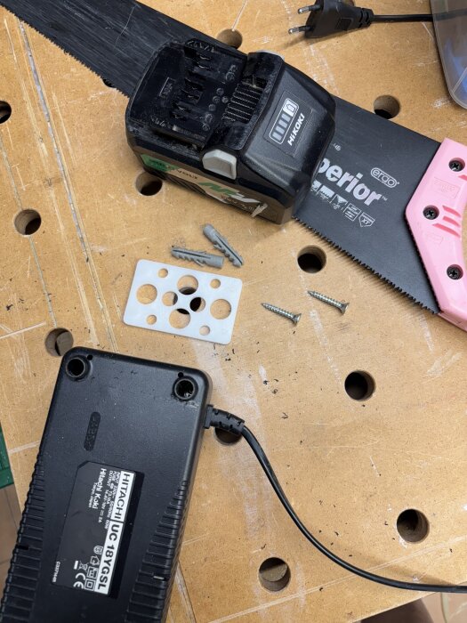 Verktyg och skruvar på ett arbetsbord, inklusive en såg, plugg och laddare.