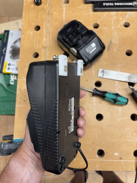 En hand håller ett verktyg med monterade fästen, placerat på en arbetsbänk med skruvmejsel och laddare i bakgrunden.