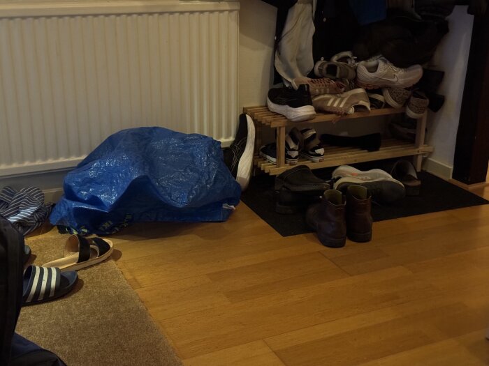 Skoställ med skor i olika stilar och en blå plastväska liggande på ett trägolv nära ett element.