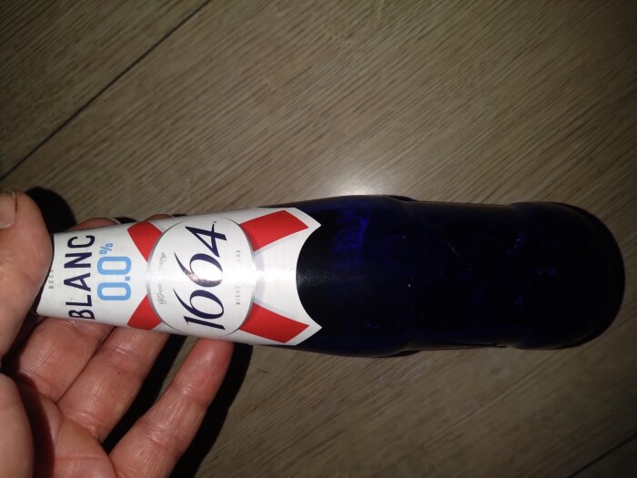 En hand håller i en blå glasflaska av alkoholfri 1664 Blanc öl på ett träbord.