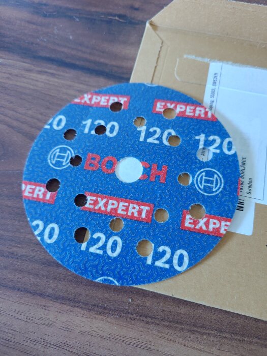 Slipskiva av märket Bosch Expert med 120 grit, liggande på ett bord och delvis ovanpå ett papperskuvert.