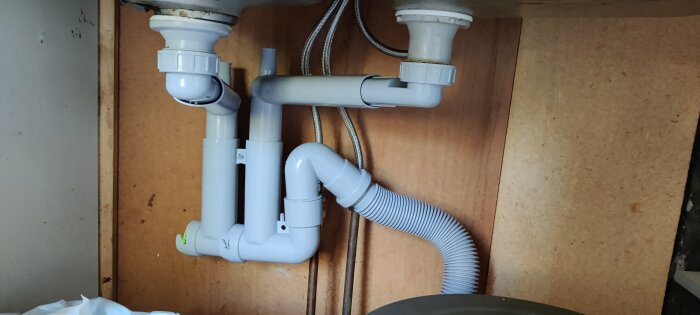Rördragning under diskbänk med vita plastavloppsrör och silverfärgade flexibla kopplingsslangar.