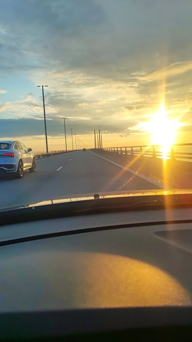 Solnedgång över en bro med bilar körande i båda riktningarna, sett från en bils vindruta.