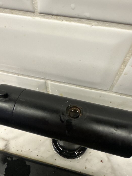 Svart köksblandare med en synlig skruv på toppen, delvis lossad, mot en vit kakelvägg.