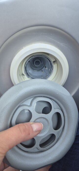 Användare håller i en rund plastdel av en spabaddel. Bakgrunden visar en del av spabadet där delen ska monteras.