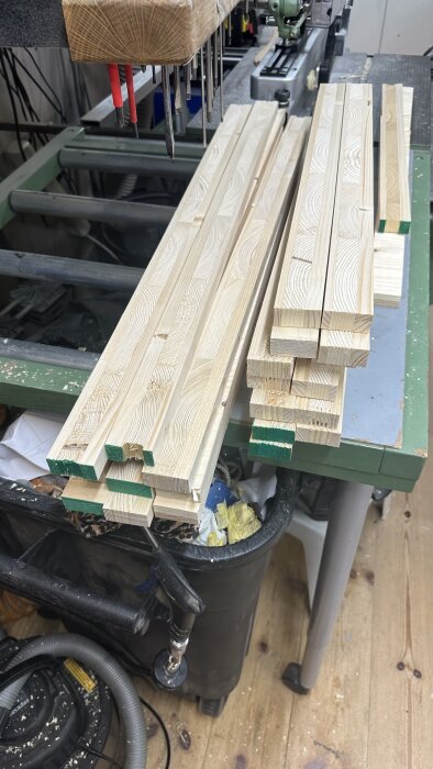 Staplade träbitar med grönmärkta ändar på en arbetsbänk i en verkstad, omgivna av verktyg och utrustning.