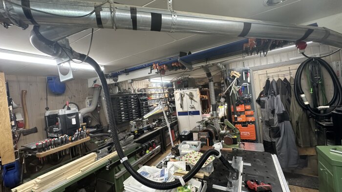 Verkstad med verktyg och utrustning inklusive sugportar, fräsbord och kappsåg; arbetskläder hänger på väggen och olika trädelar ligger på bordet.