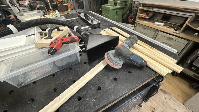 Arbetsbänk med elverktyg, sugslang och träbitar, omgiven av verktygslådor och snickeriverktyg i en verkstad.