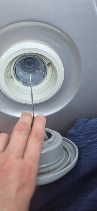 Hand avlägsnar en rund, grå avloppsdel från en tvättmaskin med ett verktyg, omgiven av en blå trasa.