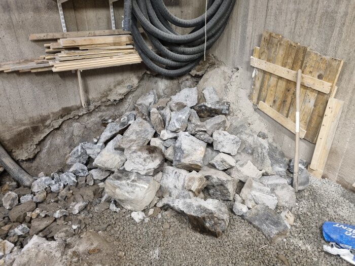 En hög med stenar vid en vägg, omgiven av byggmaterial och rör, i ett pågående byggprojekt.