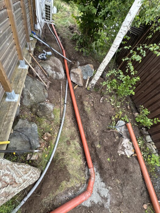 Rött avloppsrör med snäv böj vid fritidshus, omgivet av jord och stenar.