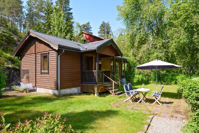Ett 1-plans trähus med veranda, omgiven av grönska. Framför huset står ett trädgårdsmöblemang med parasoll.